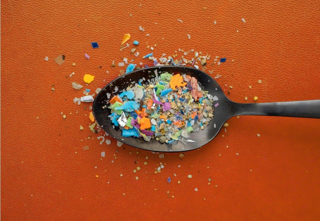 A dark spoon holds colorful microplastic fragments and granular debris on an orange background.